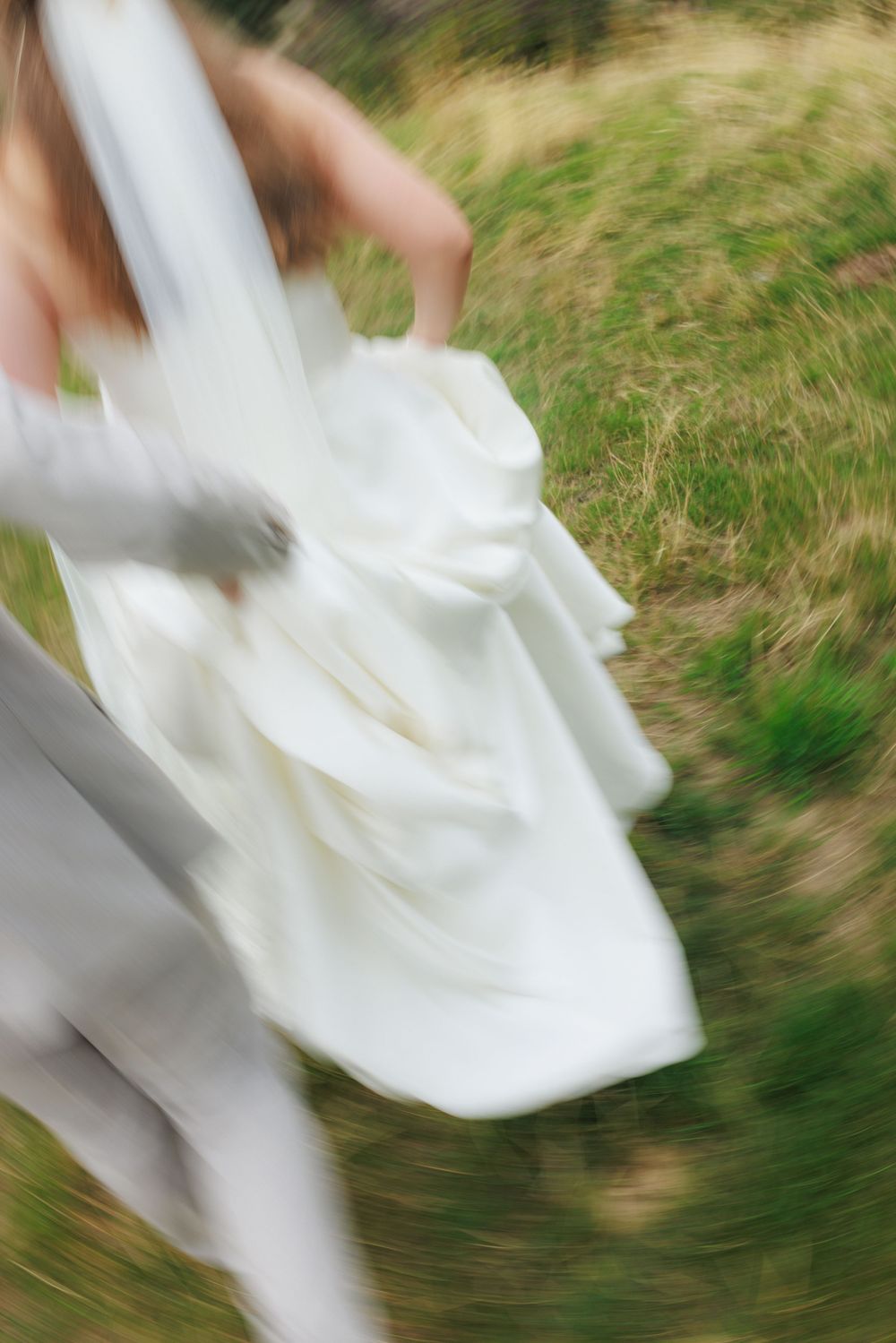Blurred motion shot of a bride in a white wedding dress and veil running through a green grassy field.