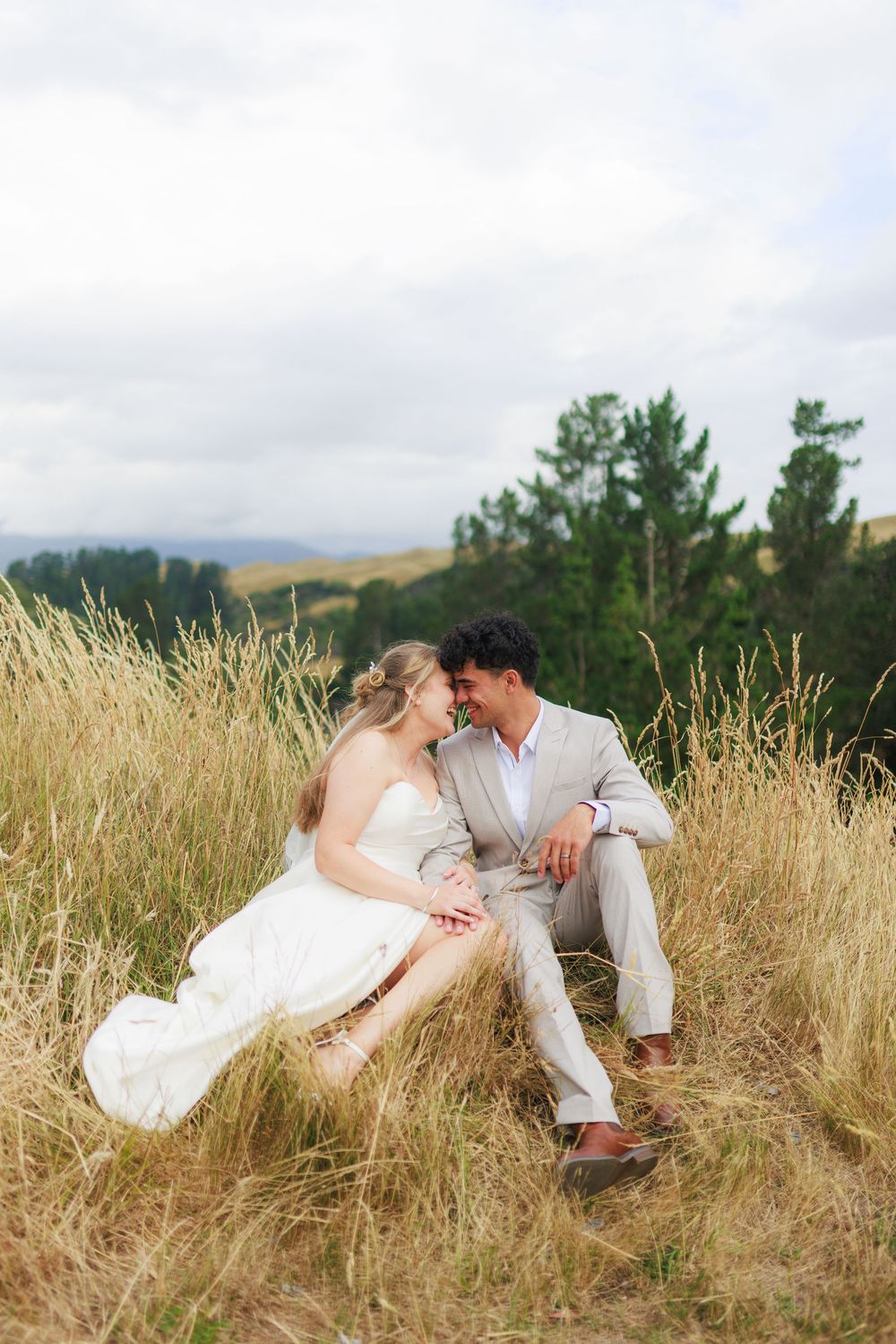 Bride and groom sitting together in tall golden grass field, sharing a romantic moment with trees and cloudy sky behind them.