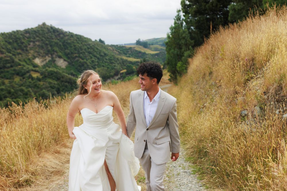 Bride and groom walking hand in hand on a scenic hillside path surrounded by golden grass and lush green mountains.