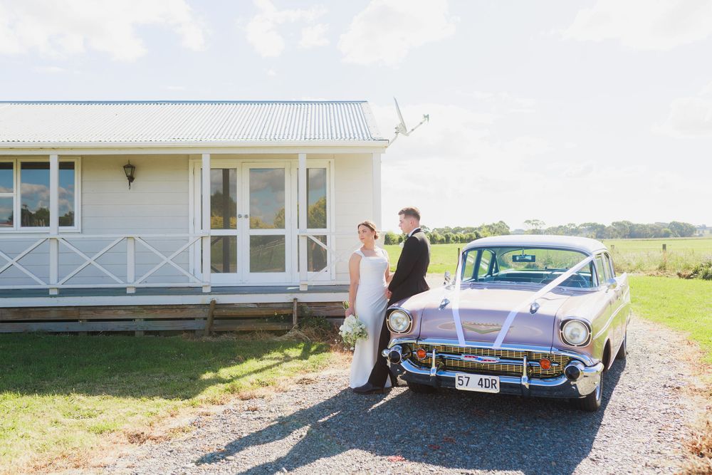 Bride and groom posing beside a vintage classic car outside a white country homestead on their wedding day.
