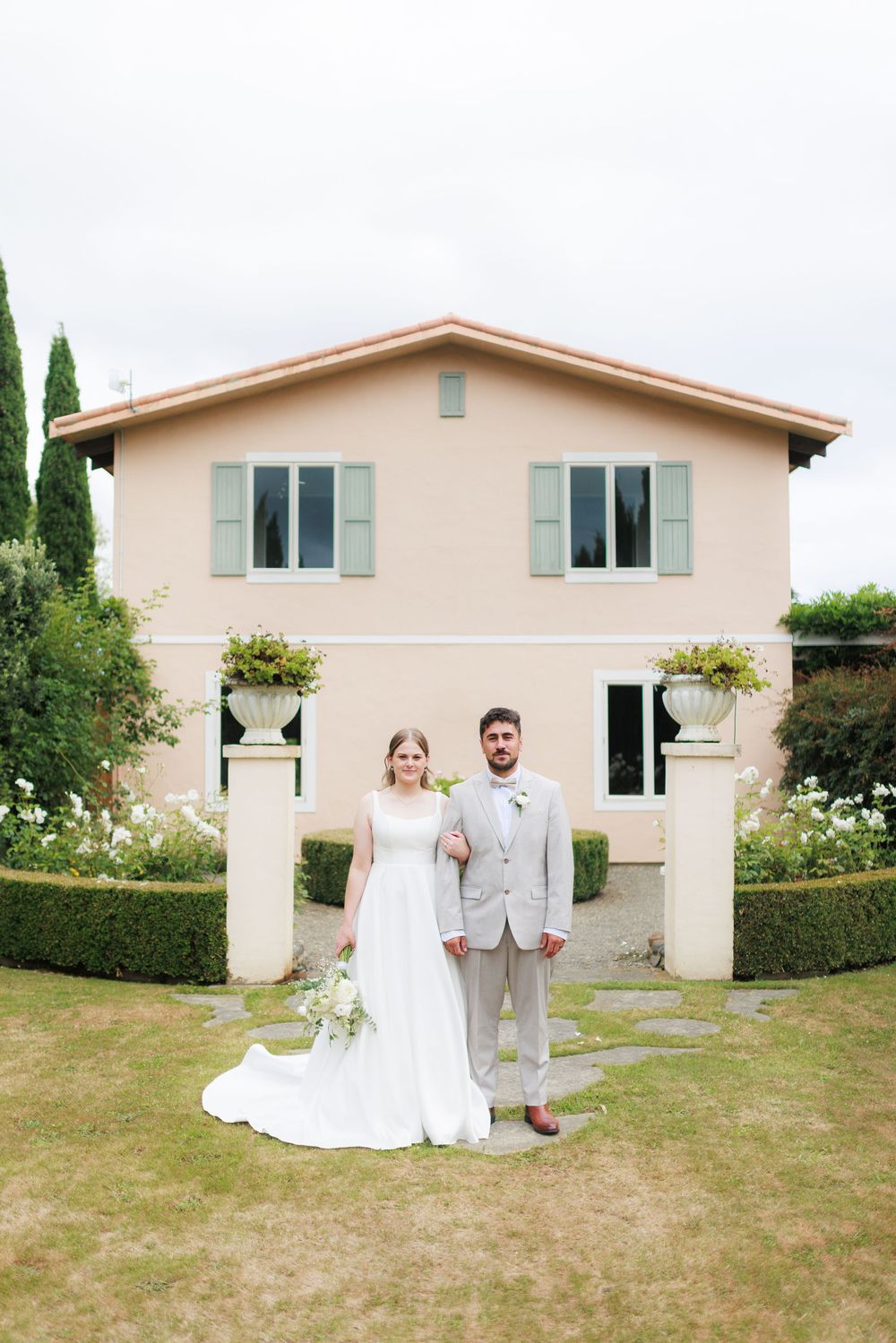 Bride and groom pose in front of a pink villa with green shutters and manicured gardens on their wedding day.