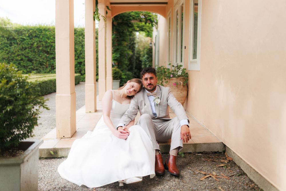 A bride and groom sit together on steps outside a elegant villa, she in white gown, he in gray suit with brown boots.