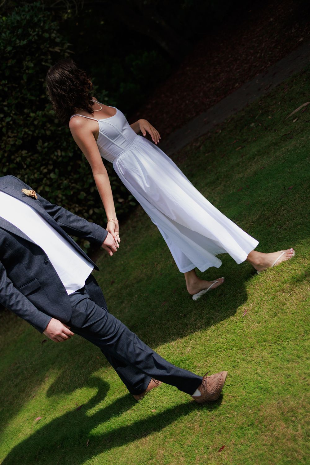 Bride in white dress and groom in dark suit walking hand in hand on green grass outdoors.