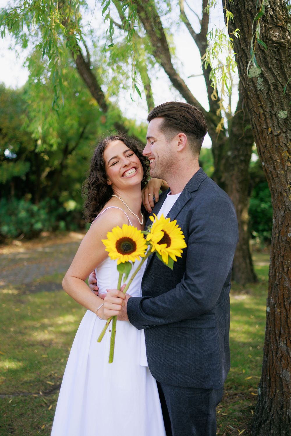 A smiling bride in white dress holds sunflowers while groom in gray suit embraces her under a tree outdoors.