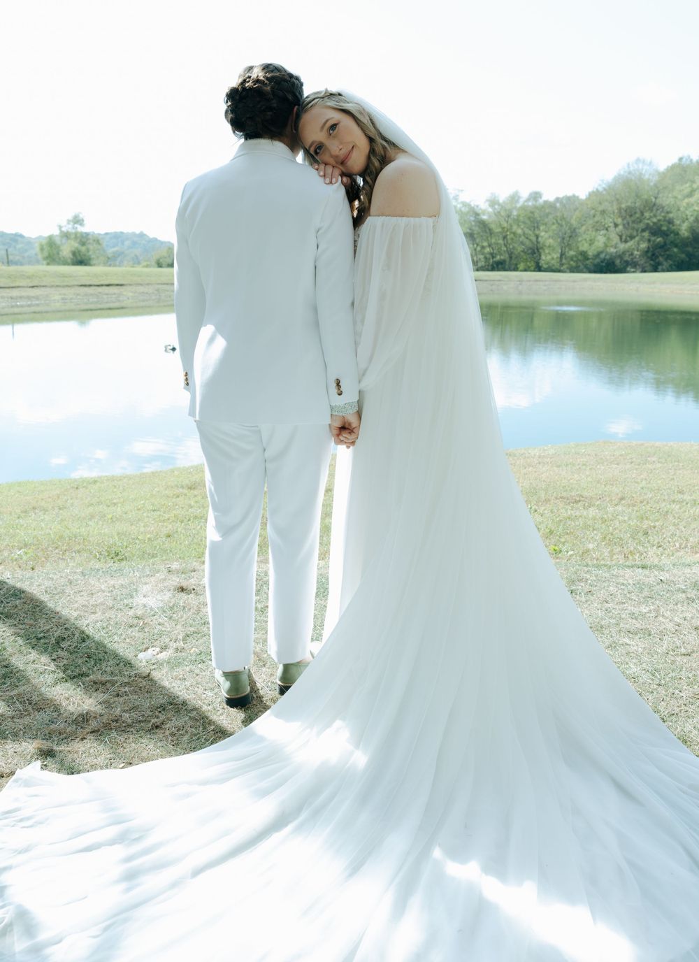 A couple in all white wedding attire stands together by a serene lake with flowing gowns caught in soft sunlight.