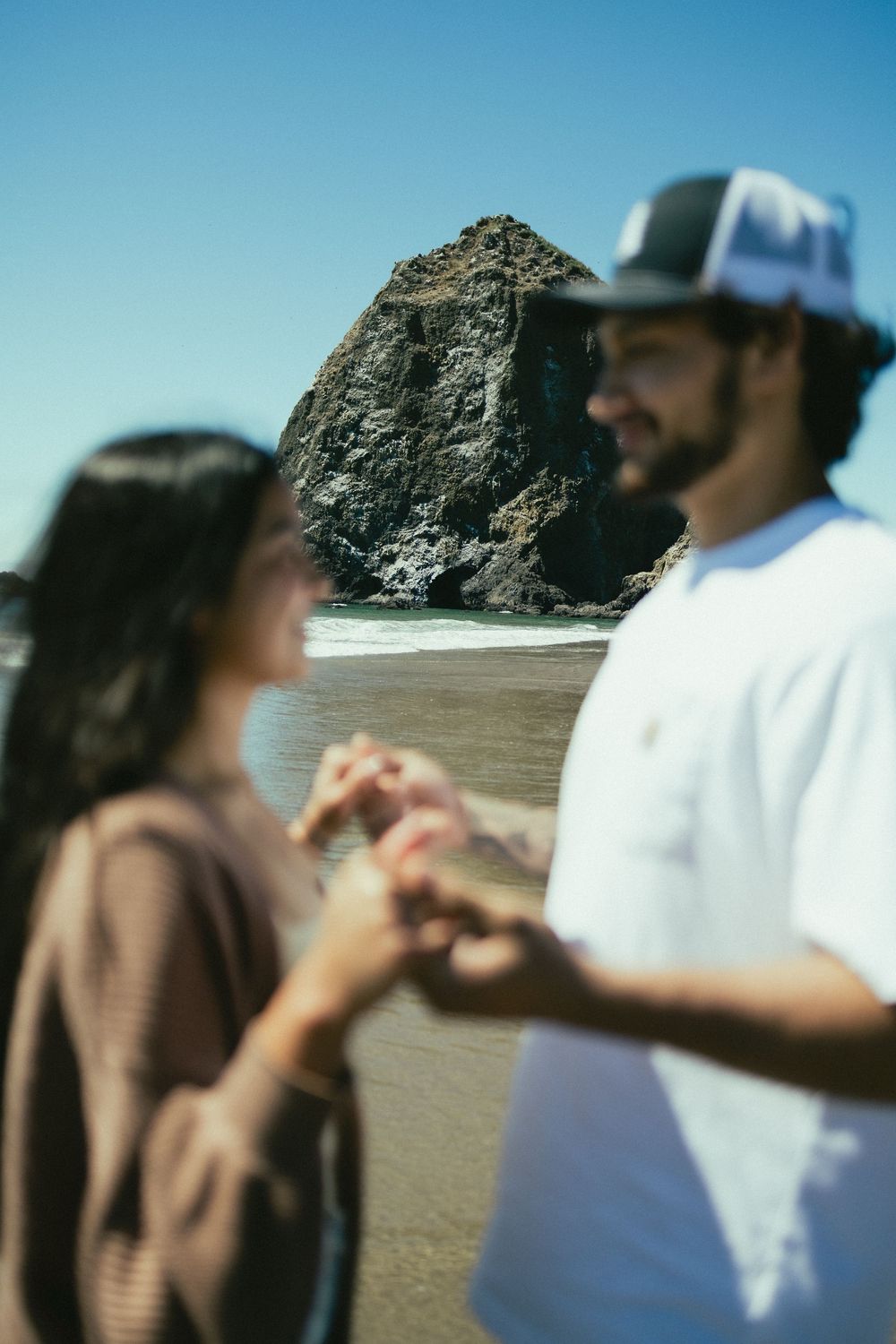 A couple holds hands on a beach with a large rocky formation visible in the background against a blue sky.