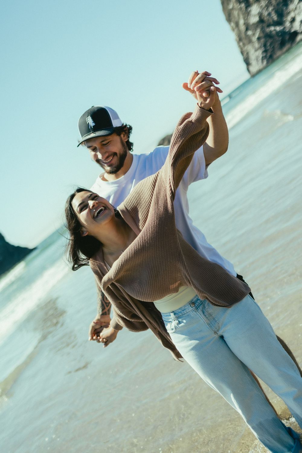 A couple dances playfully together on a sunny beach with ocean and cliffs in the background.