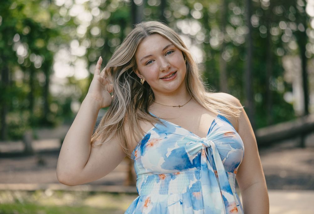A person in a light blue and white floral dress poses outdoors in a park setting during golden hour lighting.