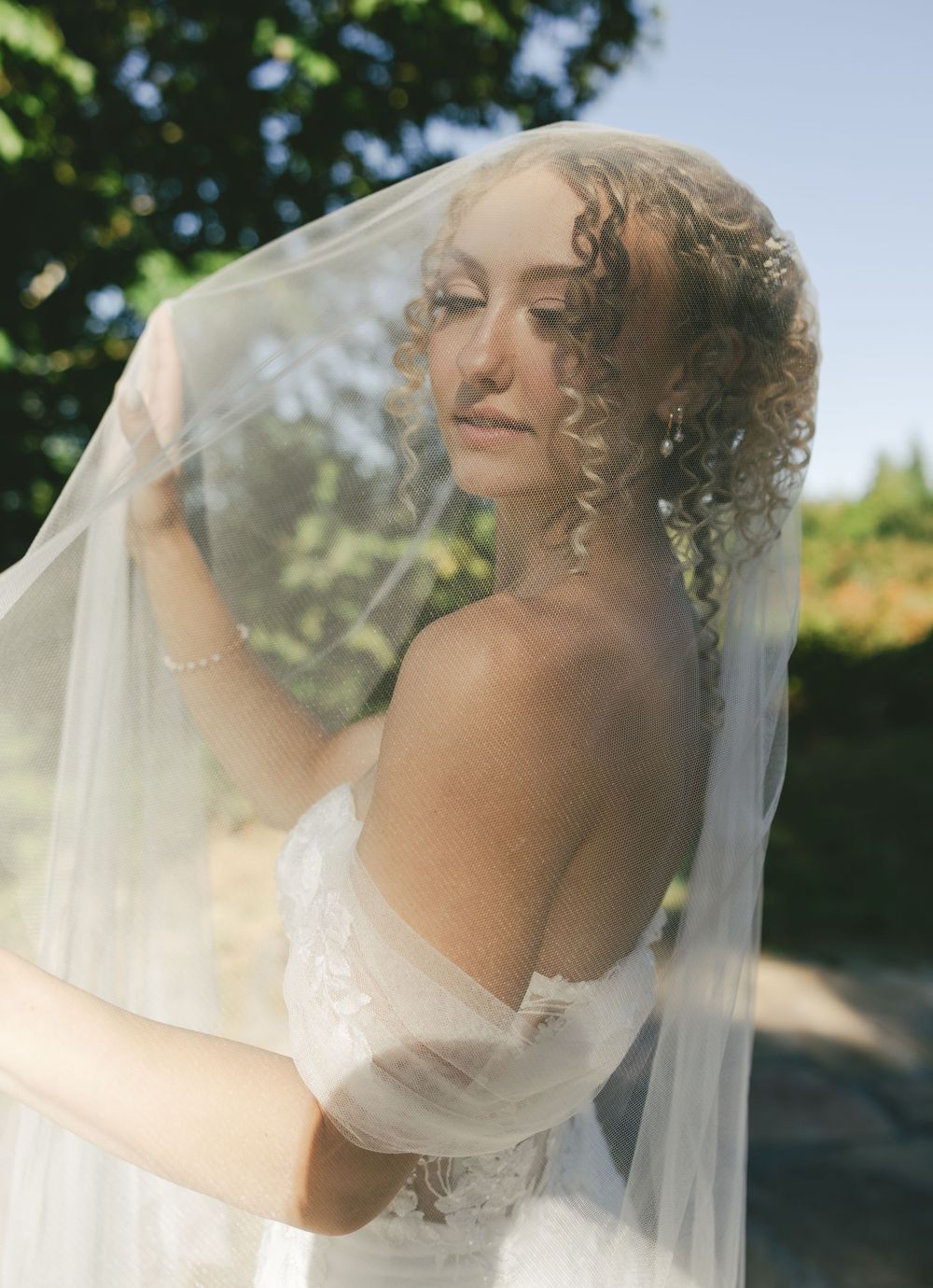 A bride in a flowing white wedding dress and veil poses outdoors on a sunny day with trees in the background.