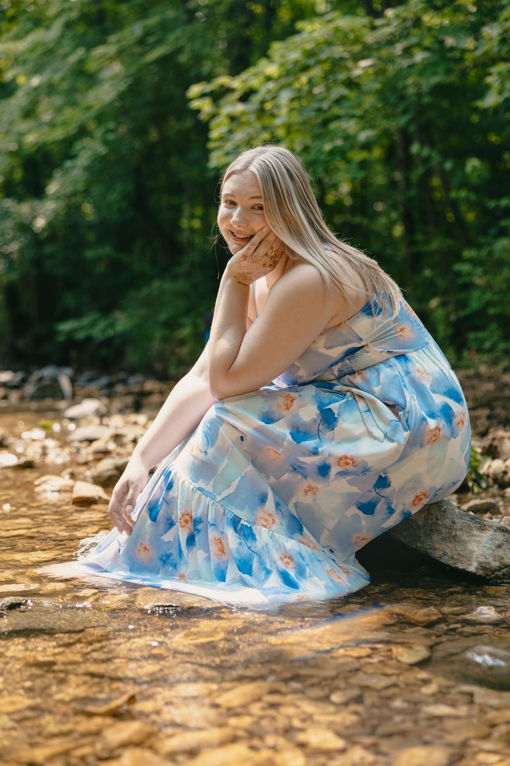 A person in a flowing blue floral dress crouches by a stream in a lush forest setting.