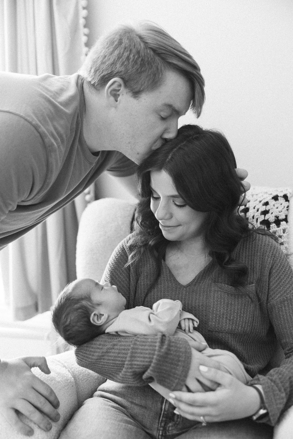A touching black and white family portrait showing parents lovingly gazing at their newborn baby in a tender moment at home.