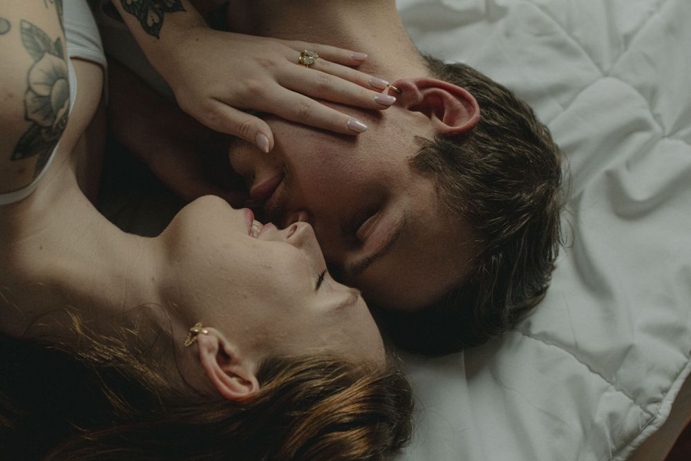Two people share an intimate moment lying close together on white bedding in moody lighting.