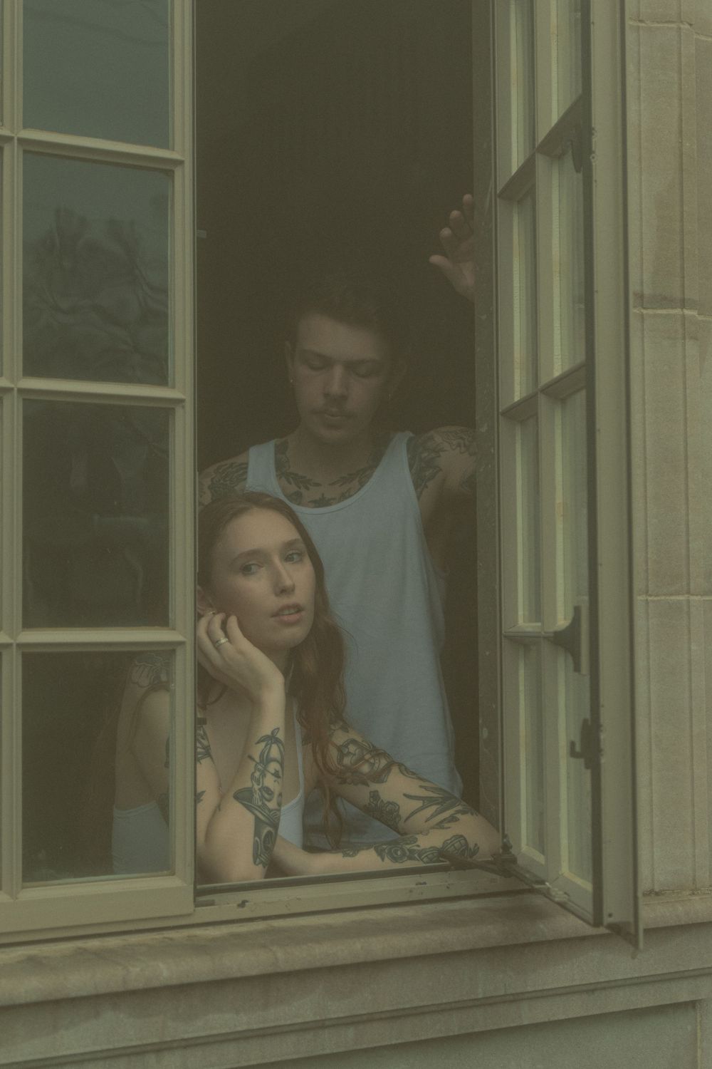 Two people gaze pensively through an open window in a moody, atmospheric portrait.