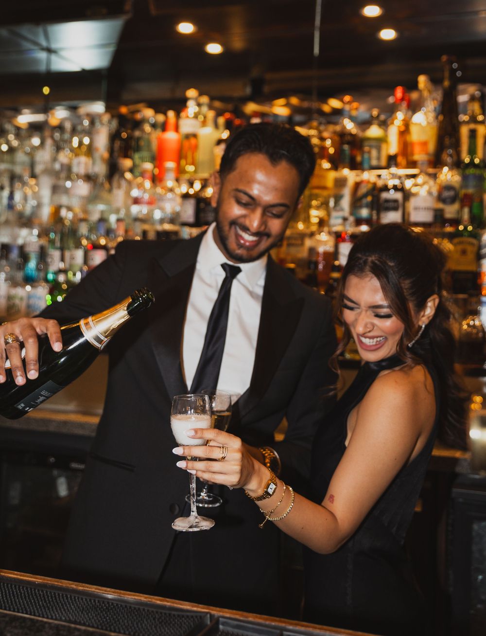 Couple sharing drinks and laughter at an elegantly lit bar in formal attire.