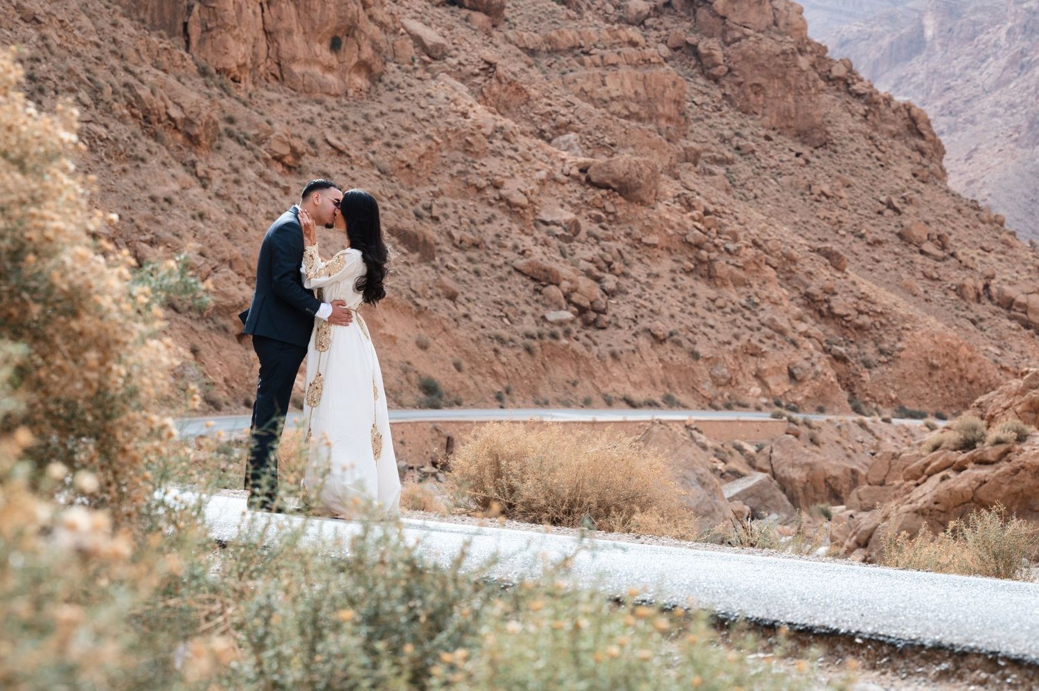 Two people in wedding attire embrace on a mountain road with dramatic red rock cliffs in the background.
