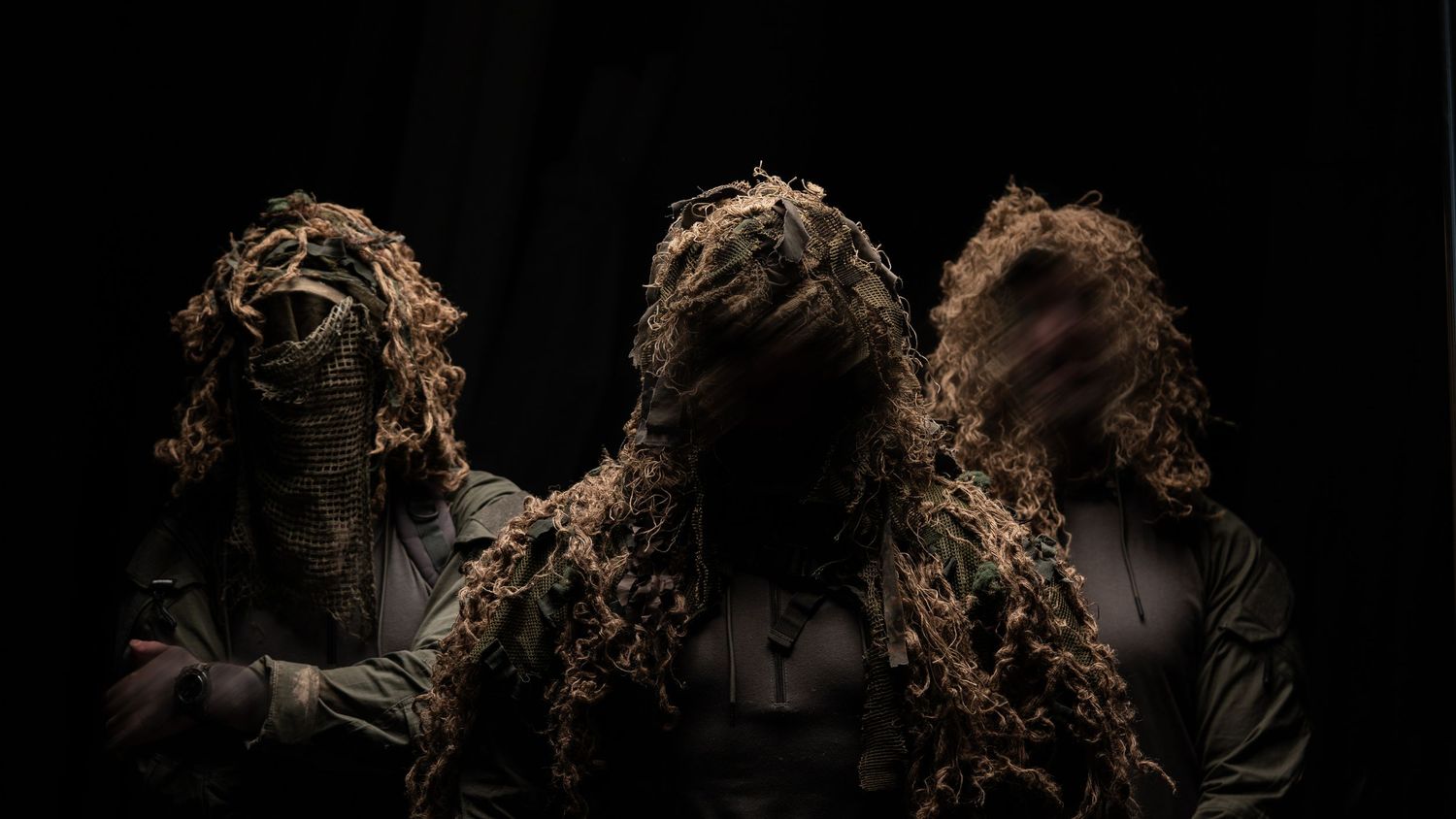 Three figures in ghillie suits pose against dark background creating dramatic silhouette effect.