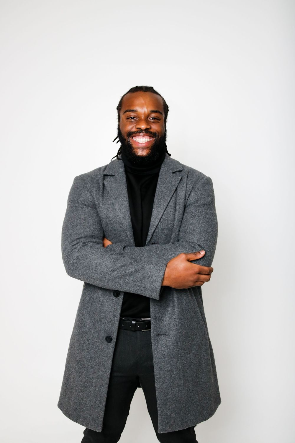 Person in grey coat and black turtleneck smiles warmly against white backdrop.