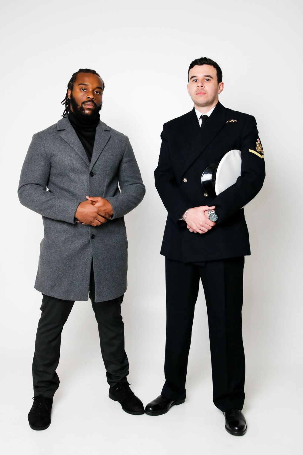 Two people in contrasting black and gray formal suits pose together against a white backdrop.