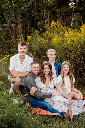 Family of five smiling outdoors on a blanket surrounded by lush green and yellow wildflowers during golden hour.