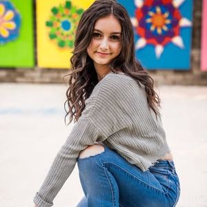 A person wearing a gray sweater and jeans poses outdoors against a colorful floral backdrop.