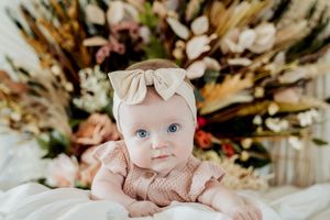 A baby in a pink outfit with a bow lies on a soft surface surrounded by autumn-toned floral arrangements.
