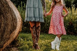 Brown leather cowboy boots peek out beneath a pink floral dress while standing in sunny green grass.