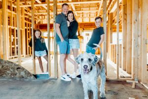 A happy dog stands with a group of people inside the wooden framing of a home under construction.