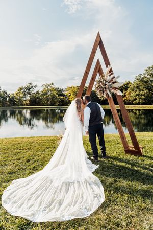 A couple in wedding attire stands before a triangular wooden arch by a serene lakeside on a sunny afternoon.