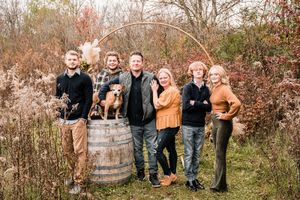 Family of five poses outdoors near a wooden barrel and circular arch, with a dog, surrounded by autumn foliage.