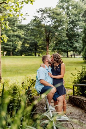 A couple shares a romantic moment together on a stone wall in a lush park setting on a sunny day.