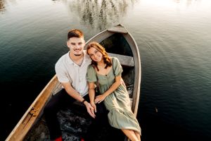 A couple sits together in a wooden rowboat on calm waters during sunset.