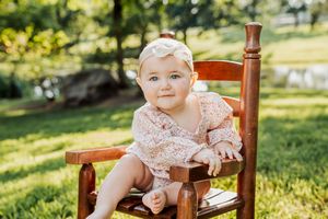 Baby in floral dress sits in wooden rocking chair outdoors on sunny lawn.