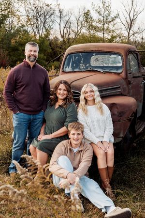 Family of four posing outdoors near a rustic vintage truck in a field during autumn.