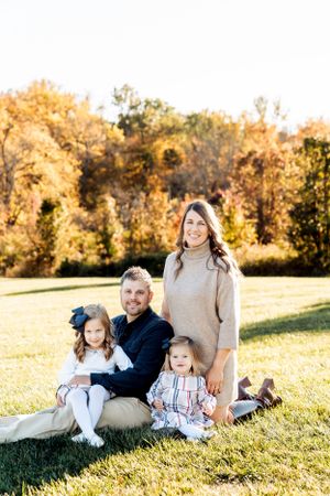 Family sitting together on grass during autumn with golden fall foliage in background on sunny day.