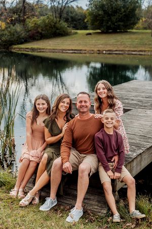 Family of five smiling together on a wooden dock beside a serene pond surrounded by autumn trees.