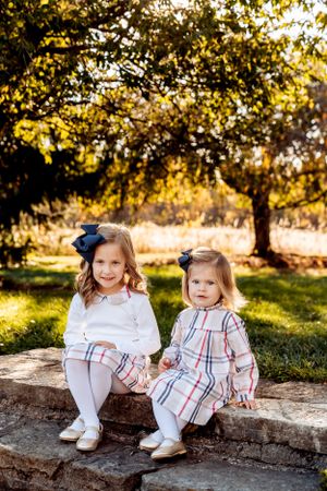 Two kids in matching plaid outfits with blue bows sit on a stone wall in a sunny autumn garden setting.