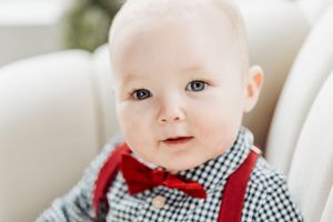 A baby wearing a red bowtie and black and white gingham shirt with suspenders looks toward the camera.