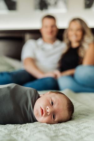 A baby lies on a textured blanket in the foreground while two people sit together on a couch in soft focus behind.