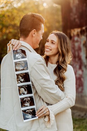 Happy couple embracing outdoors holding ultrasound photos, announcing pregnancy in warm autumn sunlight.