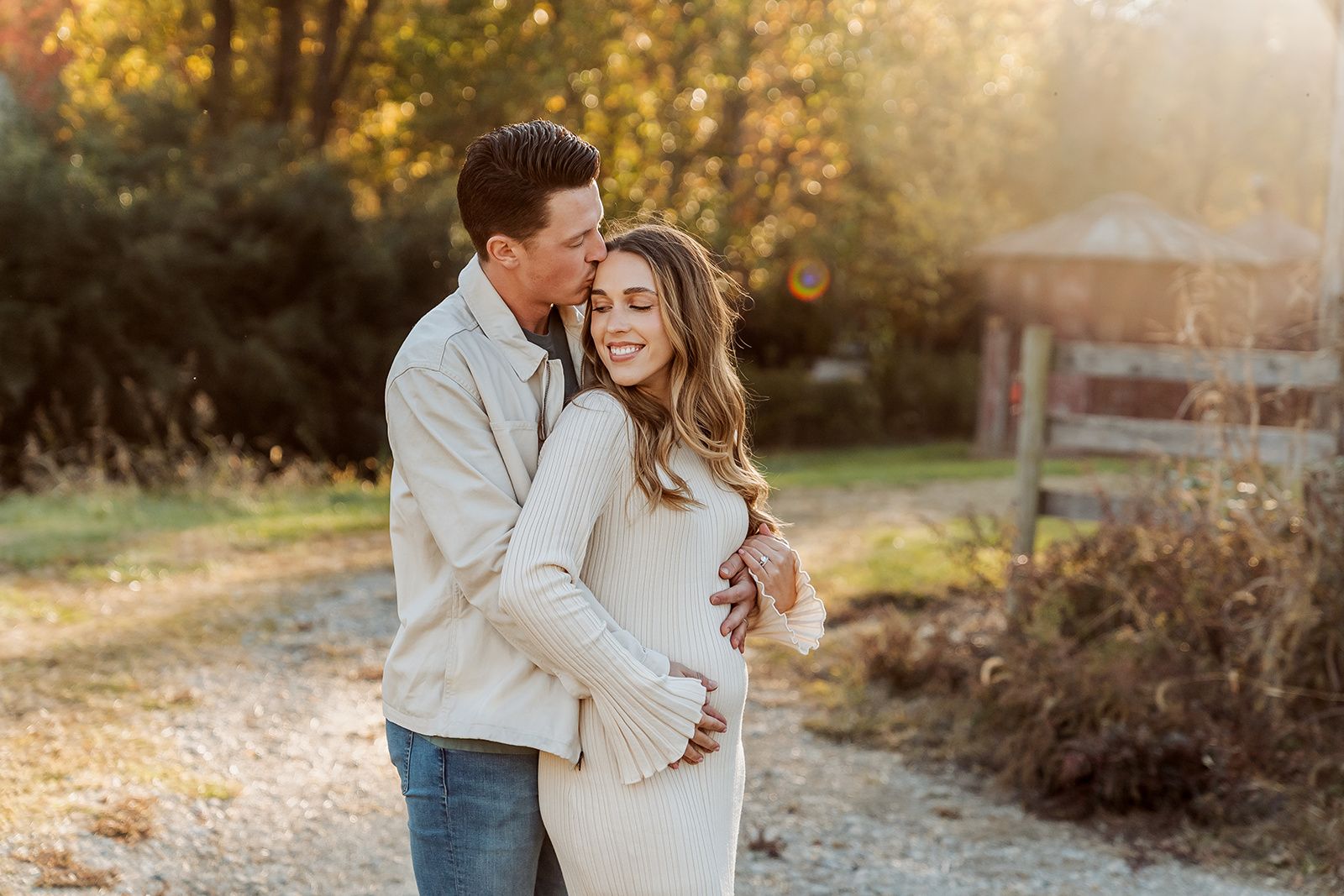 Couple embracing outdoors during golden hour, pregnant woman in white dress smiling as man kisses her cheek near wooden fence.