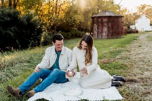 Couple sitting on a white blanket outdoors at golden hour, laughing together near a rustic barn in a green field.