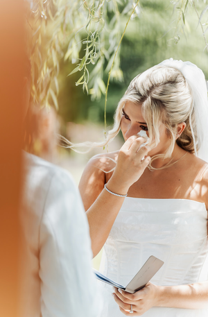 Emotional bride in white strapless gown wiping tears during outdoor wedding ceremony under willow tree.