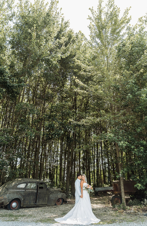Bride and groom embrace before tall trees and a vintage rusty car at an outdoor rustic wedding venue.