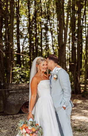 A bride in a white strapless gown holds a colorful bouquet as a groom in a light blue suit whispers in her ear outdoors.