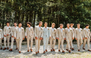 Groom and groomsmen in matching tan suits with brown shoes standing outdoors in front of lush green trees.