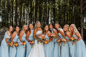 Bride in white gown with nine bridesmaids in light blue dresses holding colorful bouquets outdoors.