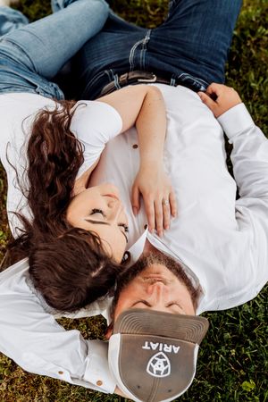 Young couple lying on grass wearing white shirts and jeans, with their heads close together and hair spread out.