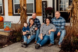 A group wearing coordinating plaid shirts sits on a porch decorated with autumn cornstalks and pumpkins.