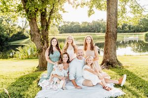 Family of six poses outdoors on blanket by pond under trees in golden hour light.