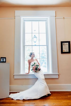 Bride in strapless white mermaid gown holding pink bouquet, posing by tall window with natural light on hardwood floors.